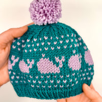 A hand-knit teal and lavender baby hat featuring a whale colorwork motif and a purple pom-pom.