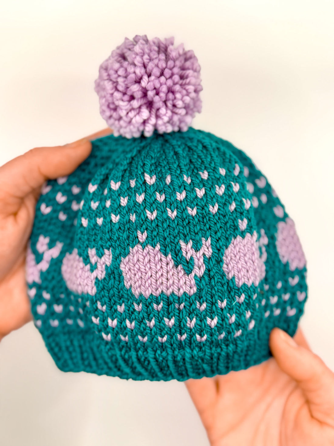 A hand-knit teal and lavender baby hat featuring a whale colorwork motif and a purple pom-pom.